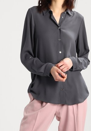 Vrouw draagt een donkergrijs, lang mouw knoophemd en lichtroze broek, staand met handen licht samen geklemd voor zich.