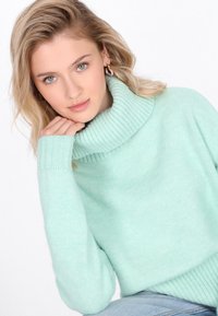 Svijetlozelena turtleneck pletiva od mekog materijala s rebrastim rukavima i ovratnikom. Dizajn uključuje opuštene rukave i uski rub.