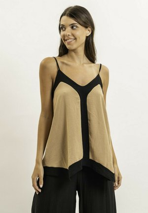 Femme portant un haut ample sans manches beige et noir à fines bretelles, associé à un pantalon noir, debout devant un fond clair uni.