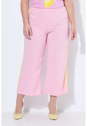 Vrouw die roze broek met wijde pijpen en gele zijstrepen draagt en gele puntige hakken, staand tegen een witte achtergrond.