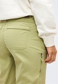 Άτομο που φοράει ανοιχτό πράσινο παντελόνι με πίσω τσέπη με καπάκι και πλαϊνή τσέπη με φερμουάρ, σε συνδυασμό με λευκό μακρυμάνικο μπλουζάκι.