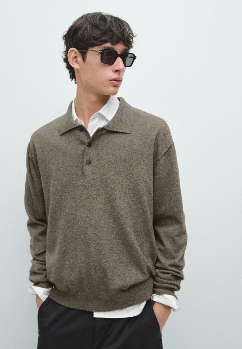 Brauner Strickpullover mit Kragen, drei Knöpfen und lockerem Schnitt. Wird über einem weißen Hemd mit Knöpfen getragen. Schwarze Sonnenbrille vervollständigt den Look.