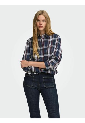 Jeune femme aux longs cheveux blonds portant une chemise à carreaux bleu marine, blanc et rouge et un jean bleu foncé, debout, les bras croisés.