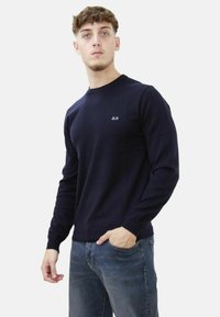Maglione lavorato a maglia blu navy con maniche lunghe, polsi a coste e scollatura tonda. Presenta un piccolo logo sul petto, abbinato a jeans blu.