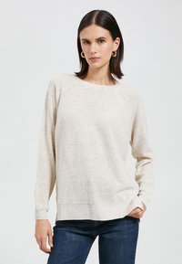 Beige gebreide trui met een ronde halslijn, lange mouwen, een gestructureerd patroon, en zijsplitten, gecombineerd met donkerblauwe jeans.