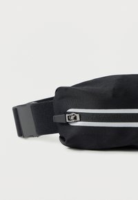 Μαύρο υφασμάτινο τσαντάκι μέσης με λεία υφή, διαθέτει φερμουάρ και λευκή ρίγα ως διακοσμητικό. Ρυθμιζόμενο μαύρο λουράκι για ασφαλή εφαρμογή.