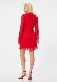Rotes, durchsichtiges Minikleid mit langen, weit ausgestellten Ärmeln und einem Rüschenausschnitt; verfügt über ein strukturiertes Muster und einen gestuften Saum. Kombiniert mit roten, geschlossenen Absatzschuhen.