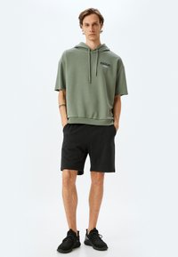 Hooded green sweatshirt with short sleeves featuring a logo, paired with black shorts and black sneakers. Simple, casual design.