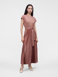 Femme portant une robe plissée mauve avec une ceinture nouée à la taille et des chaussures à talons beiges, debout devant un fond clair uni.