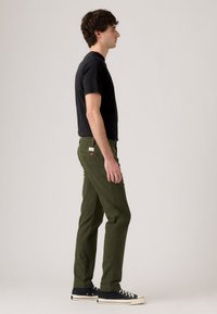 Man in black t-shirt, olive green pants, and black sneakers standing in profile against a plain light background.