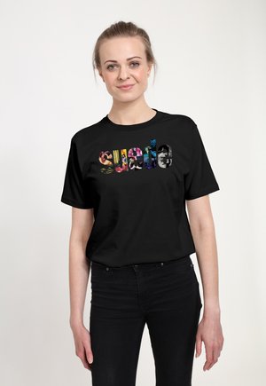 Kvinde med lys hud og blondt hår iført sort t-shirt med farverig abstrakt tekst "Sweta", stående mod en ensfarvet hvid baggrund.