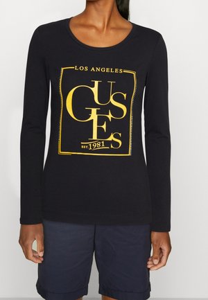 Femme portant un t-shirt noir à manches longues avec des lettres dorées "GUES", le texte "Los Angeles" et "Est. 1981", associé à un short sombre jusqu'aux genoux.