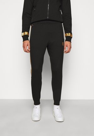 Homme portant un jogging noir avec des logos dorés EA7 Emporio Armani sur les poches et les bandes latérales, associé à des baskets blanches et des chaussettes.