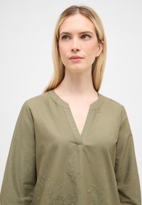 Olivengrøn skjorte med V-hals, prydet med detaljeret blomsterbroderi og lange ærmer i et glat stof. Enkel og elegant design.