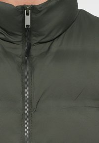 Olivovo zelená prešívaná bunda s vysokým golierom, celopresným zipsom a textúrovanou légou na zips. Hladká látka s mierne lesklým povrchom.