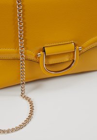 Sac à main en cuir jaune avec bandoulière chaîne dorée et détail de quincaillerie en forme de D dorée assortie sur fond blanc.