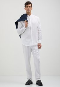 Mann in weißem Hemd und Hose, der eine marineblaue Jacke über die Schulter hält und schwarze Slipper trägt, steht vor einem schlichten hellen Hintergrund.