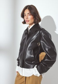 Veste en cuir noir avec fermeture éclair, large col et poche avant. Portée sur une chemise blanche et un pantalon marron, mettant en valeur une texture lisse.