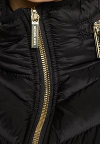 Doudoune noire avec col montant, design matelassé et fermetures éclair dorées avec tirettes de marque. Texture lisse avec une finition brillante.