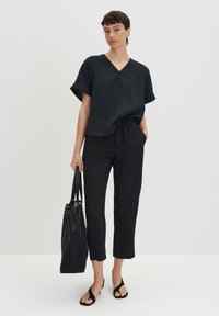 Top nero a maniche corte con scollo a V, abbinato a pantaloni neri alla caviglia. Modella tiene una borsa a tracolla nera. Sandali neri piatti.