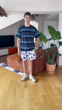 Ungdommelig mann iført en blåstripet skjorte og hvite shorts med blå mønster står i en stue med en brun sofa og en stor potteplante.