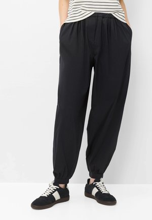 Persoon die zwarte loszittende broek draagt met elastische boorden, een zwart-wit gestreept shirt en zwarte sneakers met witte veters en zijstrepen.