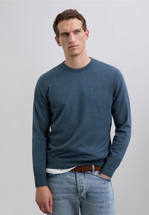 Hombre con suéter de cuello redondo azul y jeans azul claro, con cinturón trenzado marrón, de pie con una mano en el bolsillo frente a un fondo plano.