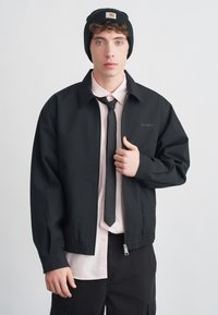 Μαύρο μπουφάν με φερμουάρ και γιακά, φορεμένο πάνω από ένα ανοιχτό ροζ πουκάμισο και μαύρη γραβάτα. Το μοντέλο φοράει μαύρο σκουφάκι με ταμπελάκι λογότυπου.