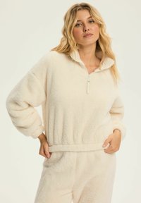 Polarowy pulower w kolorze kremowym z półzamkiem pod szyją, opadającymi ramionami oraz elastycznym paskiem w talii. Miękka tekstura, swobodny krój.