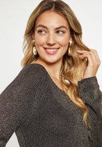 Pulover negru tricotat cu fire metalizate, având un decolteu larg și mâneci lungi. Modelul poartă cercei texturați aurii.