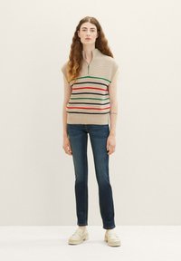 Haut en tricot beige à manches courtes, avec des rayures horizontales multicolores, fermeture éclair à l'avant, associé à un jean bleu foncé et des mocassins clairs.