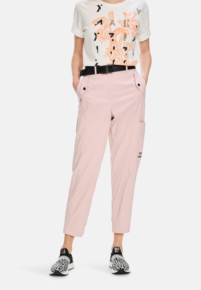 Light pink cargo pants with side pockets and black accents, paired with a white T-shirt featuring orange graphic print and rolled sleeves.