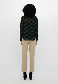 Femme aux cheveux bouclés portant une chemise noire à manches longues avec des pois verts, un pantalon beige et des bottes noires à talons, debout, de dos.