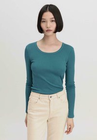 Türkises Langarmshirt mit rundem Halsausschnitt, kombiniert mit hellen, figurbetonten Hosen. Der Stoff wirkt weich und hat eine glatte Textur.