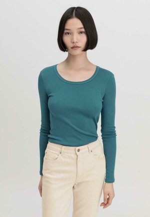 Teal longsleeve shirt met een ronde hals, gecombineerd met lichtgekleurde, aansluitende broek. De stof lijkt zacht met een gladde textuur.