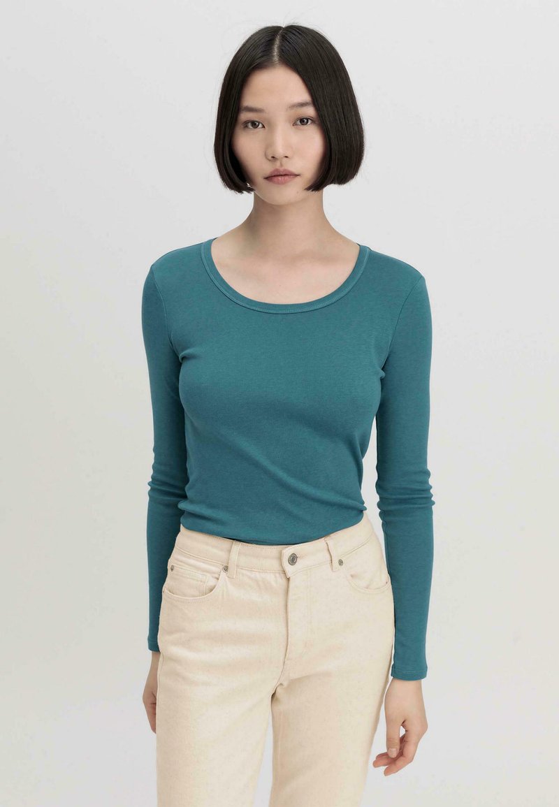 Türkises Langarmshirt mit rundem Halsausschnitt, kombiniert mit hellen, figurbetonten Hosen. Der Stoff wirkt weich und hat eine glatte Textur.
