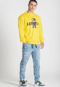 Sudadera amarilla con diseño gráfico, vaqueros rasgados de color azul claro y zapatillas coloridas con detalles de cordones. Modelo de pie contra un fondo liso.