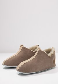 Brown suede slippers with a soft, beige fur lining, rounded shape, and contrasting stitching details along the edges. Flat soles for indoor use.