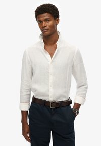 Camisa blanca de lino de manga larga con cuello alzado, frente abotonado y mangas con puños. Combinada con pantalones oscuros y un cinturón marrón.