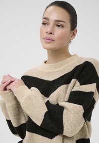 Beżowy sweter z czarnymi pasami w kształcie zygzaka, ryżowy dekolt i mankiety, miękka faktura, długie rękawy. Ręce modelki są złożone na przodzie.