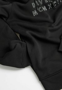 Czarna bluza z długimi rękawami, ryglowymi mankietami i okrągłym dekoltem. Z przodu znajduje się biała grafika tekstowa "DIVERSITY MCM XC".