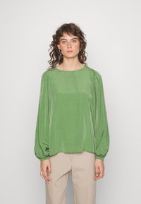 Blusa verde texturizada com mangas balão longas e um decote redondo largo, combinada com calças bege claras, contra um fundo neutro.