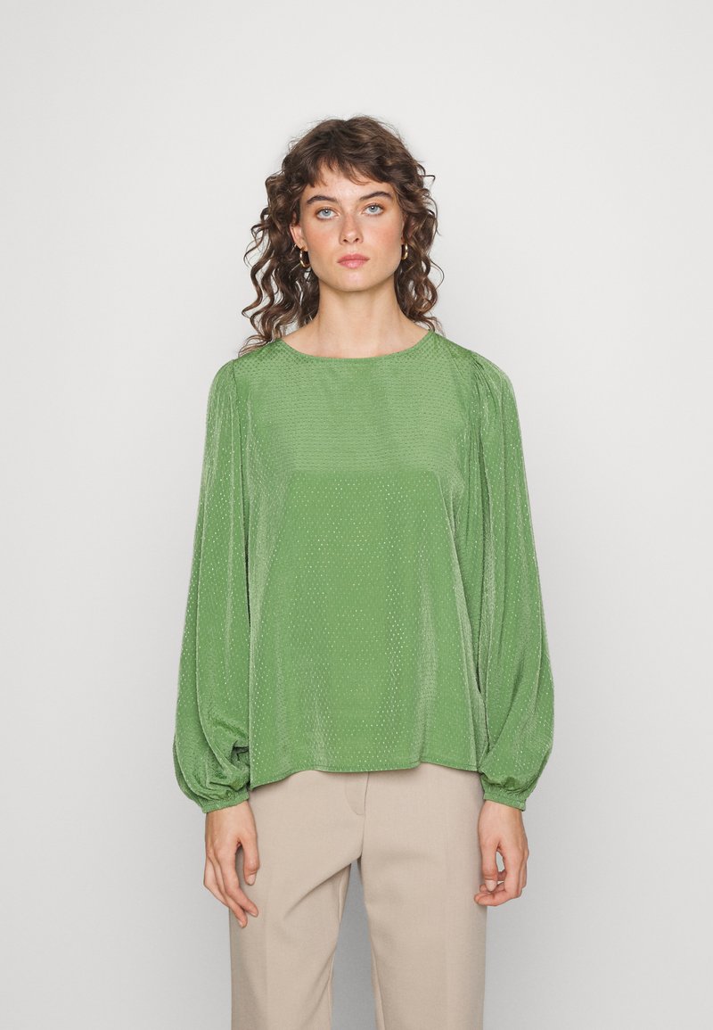 Blusa verde texturizada com mangas balão longas e um decote redondo largo, combinada com calças bege claras, contra um fundo neutro.
