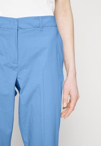 Personne portant un pantalon bleu clair taille haute et un top blanc, visible de la taille à la main sur un fond clair uni.