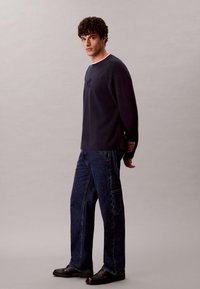 Felpa Navy con scollo a girocollo e logo ricamato, abbinata a jeans blu scuro a vestibilità larga e scarpe nere in pelle. Design semplice e casual.