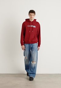 Red hoodie with drawstrings and logo, paired with light blue distressed jeans featuring frayed edges and rips. Worn with black footwear.