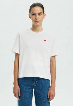 Weißes Baumwoll-T-Shirt mit kurzen Ärmeln, rundem Halsauschnitt und einer kleinen roten Herzstickerei auf der linken Seite, kombiniert mit blauen Jeans.