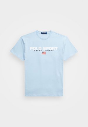 Gaiši zilas kokvilnas T-krekls ar apļa kakla izgriezumu, uz kura rakstīts "POLO SPORT" un "RALPH LAUREN" baltā tekstā, kā arī neliela Amerikas karoga grafika.