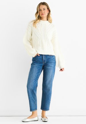 Cremfarbener Pullover mit Zopfmuster und geripptem Saum, kombiniert mit blauen geraden Jeans und silbernen Ballerinas.