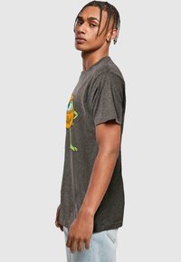 Grå t-shirt med rund halsudskæring, der har et cartoon-monster grafik i orange og grøn på siden. Stoffet ser blødt og struktureret ud.